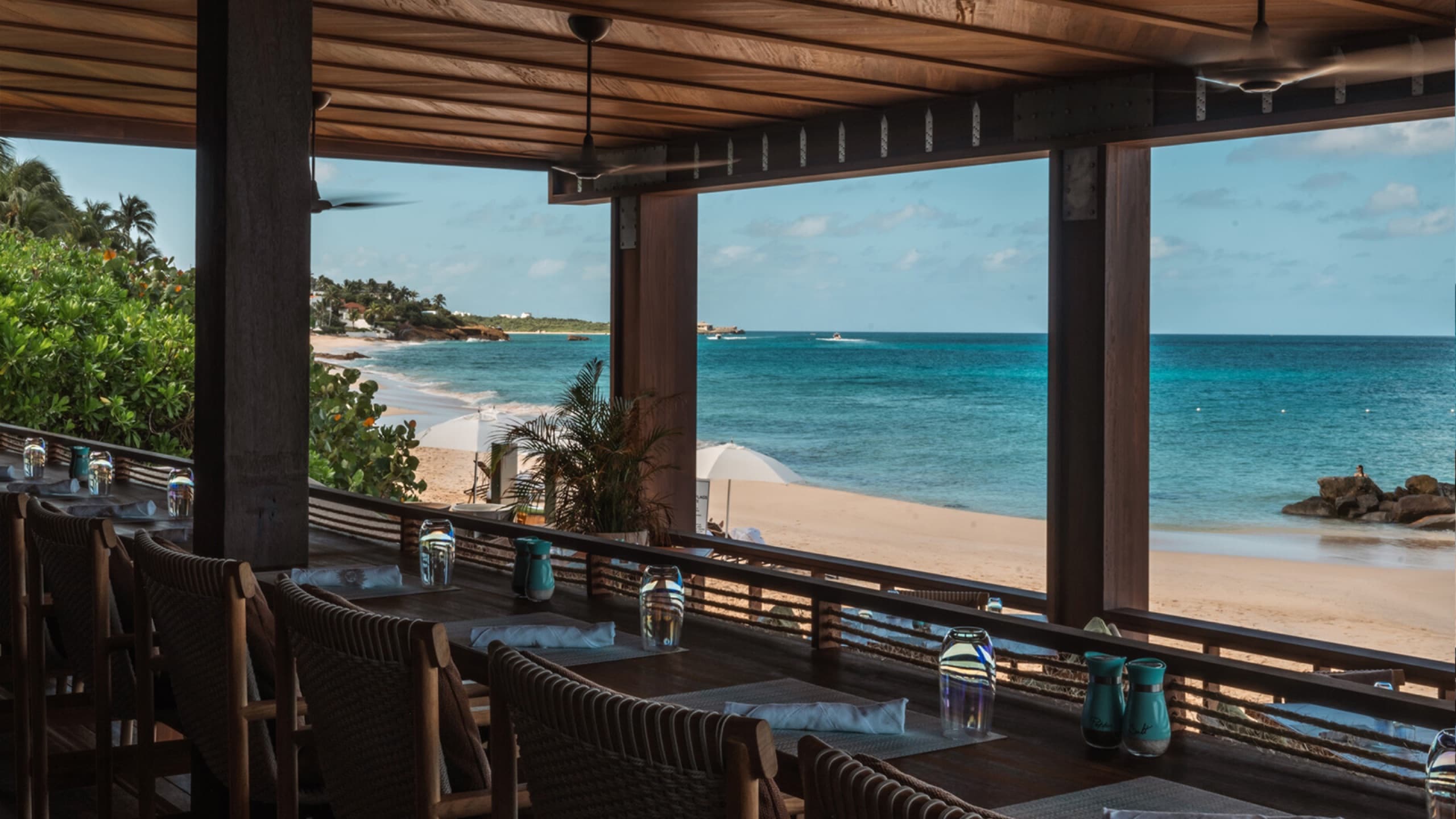 Open-air beachfront dining terrace with rattan chairs and ceiling fans, beach beyond
