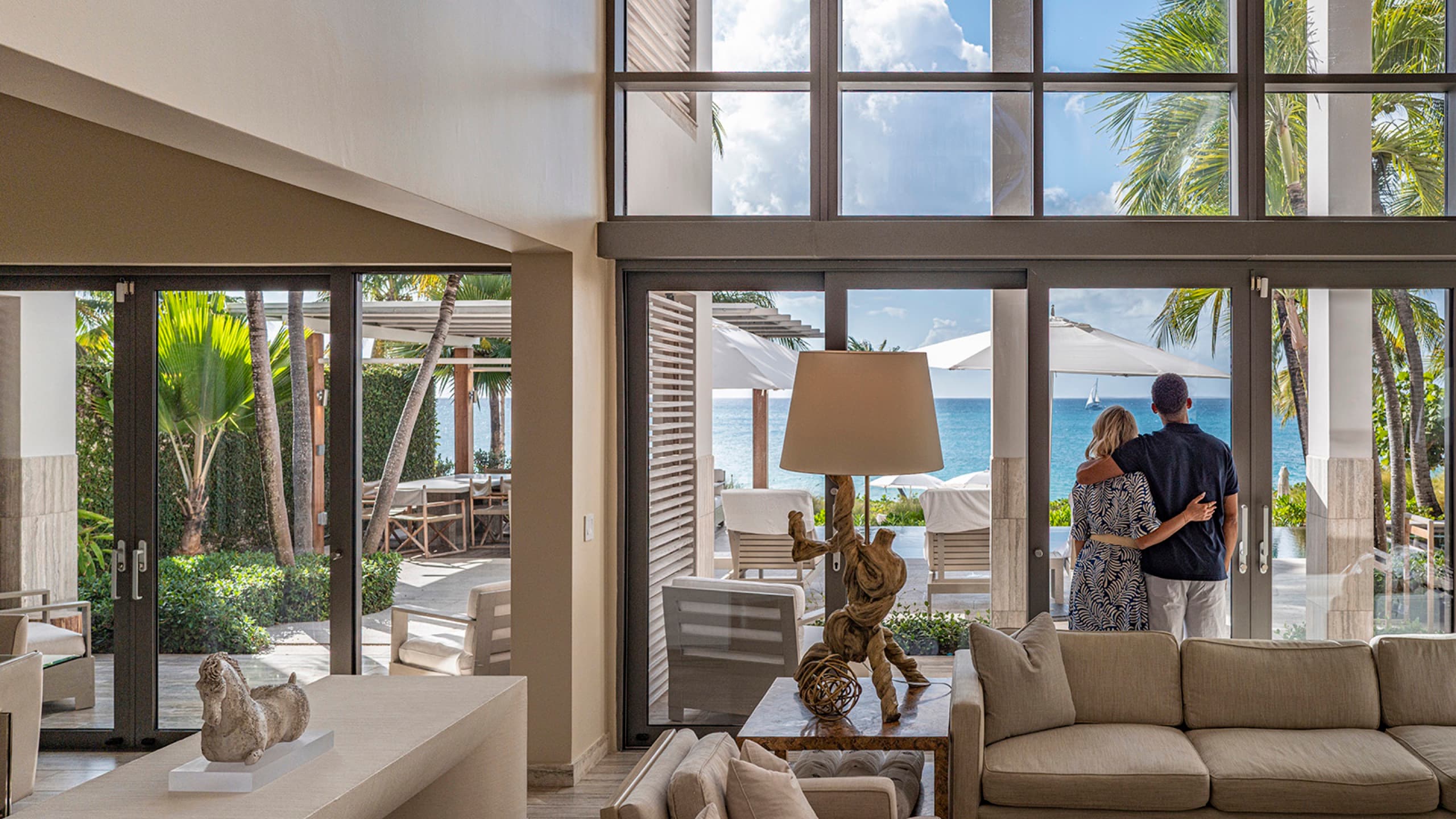 Villa living room: double-height glass wall to the terrace, sofa, driftwood lamp, sea beyond