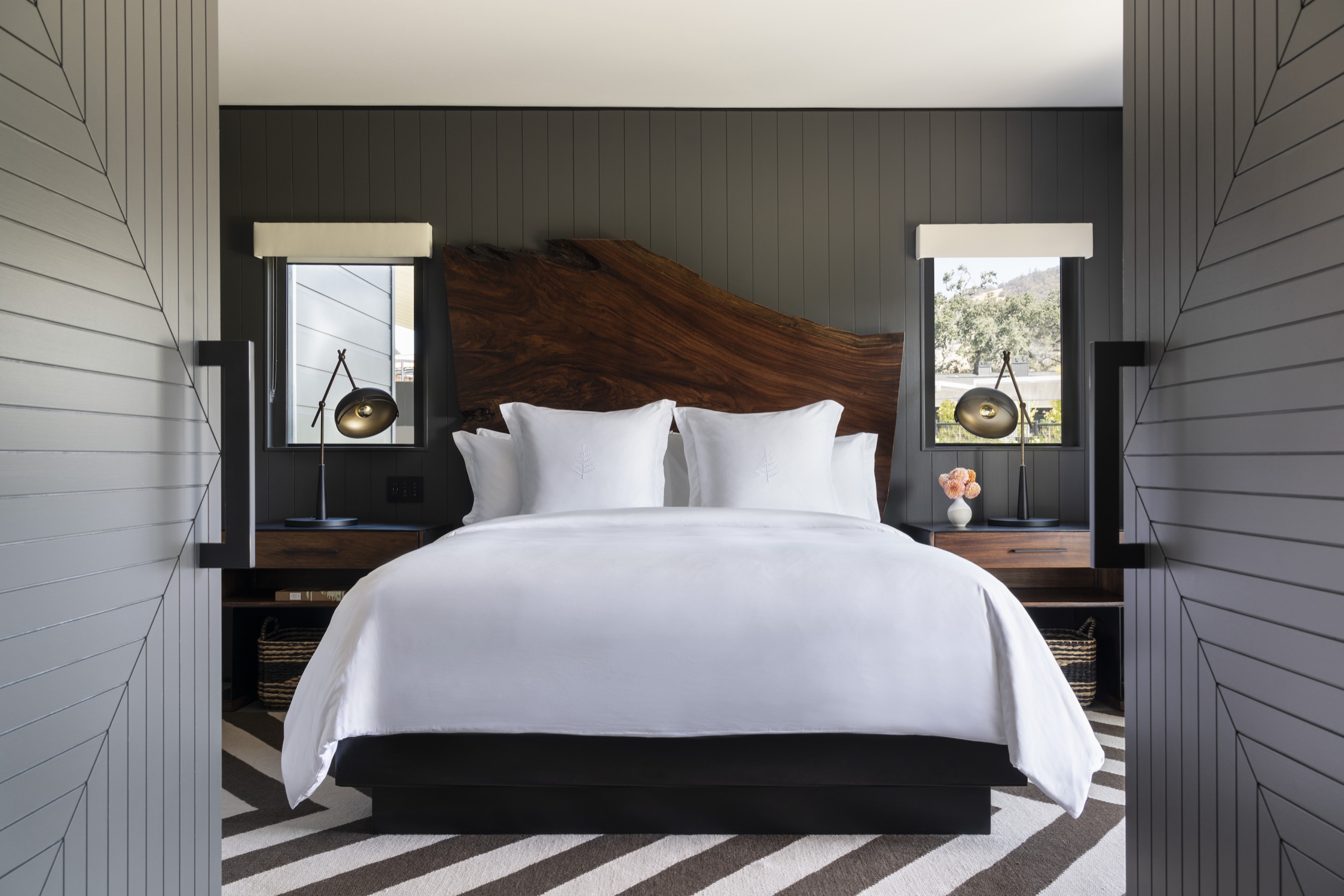 One-Bedroom Suite bedroom — king bed under a live-edge walnut headboard, black-and-white striped rug and paired windows to the vines